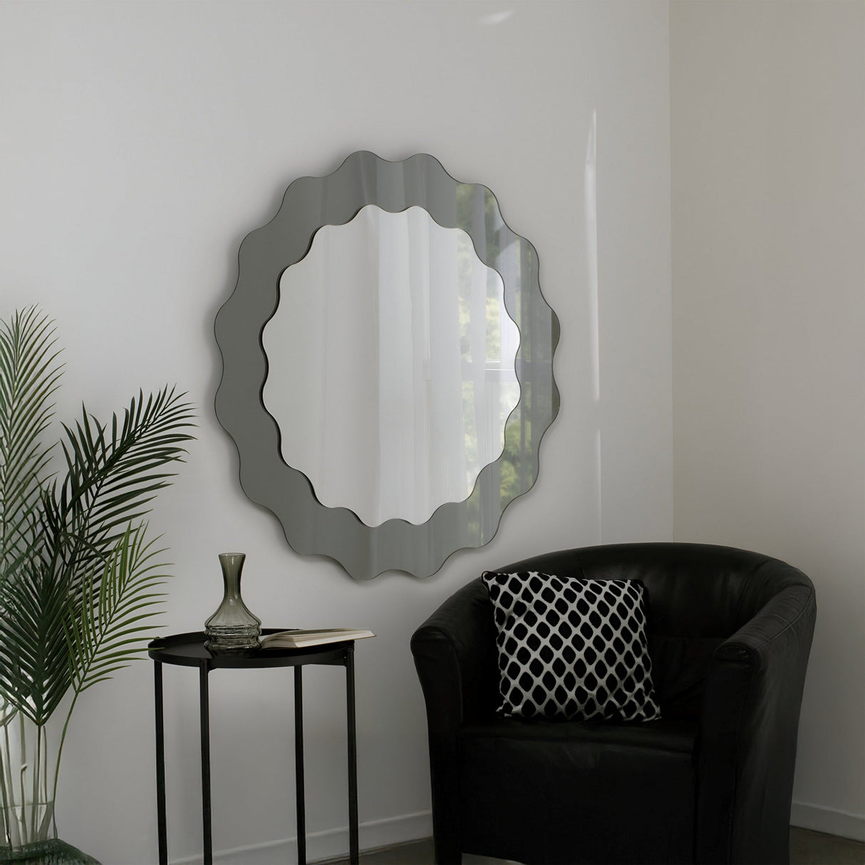 A modern living room corner featuring sculptural artistry: the Solstice Circle Wall Mirror - Grey on a white wall, a black armchair with a patterned cushion, a sleek side table, glass vase, and a green potted plant.