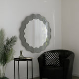 A modern living room corner featuring sculptural artistry: the Solstice Circle Wall Mirror - Grey on a white wall, a black armchair with a patterned cushion, a sleek side table, glass vase, and a green potted plant.