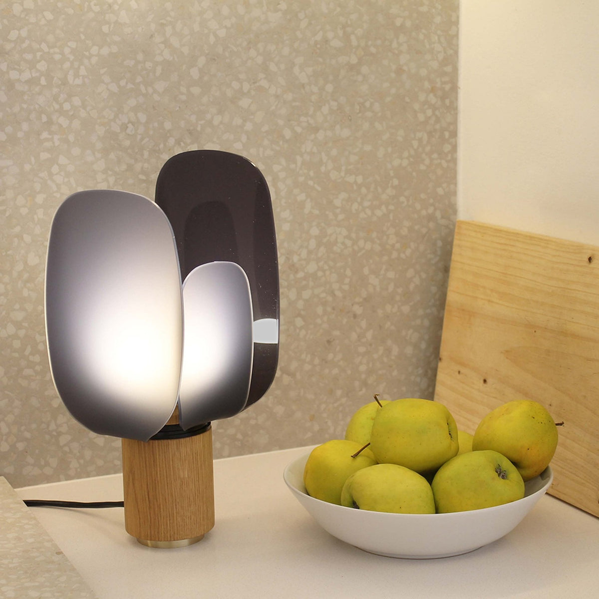 Iselmorne Wooden Table Lamp in Black, featuring curved acrylic panels on an oak base, lit beside a bowl of green apples on a kitchen counter.