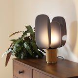Iselmorne Wooden Table Lamp in black, with curved smoked acrylic panels on a cylindrical oak base, lit atop a dark wood dresser beside a potted plant.