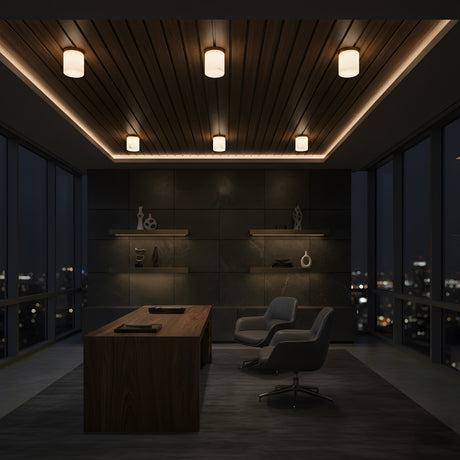 A modern office at night features a wooden desk, gray chairs, dark accent wall with shelves and decor, tall windows, and the Bravora 1 Light Cylinder Alabaster Semi-Flush Ceiling Light in Soft White & Brushed Brass illuminating the space.