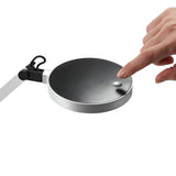 A hand touches a sleek, silver Prismora LED Table Lamp, characterized by its modern design and round black base, with an adjustable arm. The background is white.