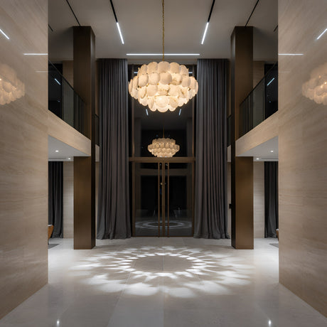 A modern, elegant lobby with marble floors, high ceilings, large gray curtains, and two Solyra 21 Light 4 Tier Petal Alabaster Chandeliers in Soft White casting intricate light patterns onto the floor.