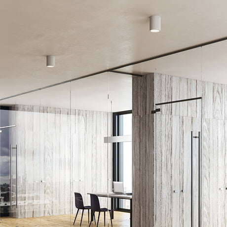 A modern office interior features wood-paneled walls and a glass partition. Inside is a minimalist desk with a laptop, a chair, and the Lucent LED Round Downlight 25W 3000K in white overhead for energy-efficient lighting. The space is bright with natural light streaming through large windows, epitomizing contemporary interiors.