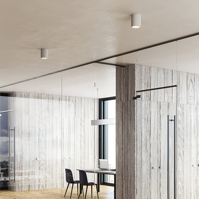 A modern office interior features wood-paneled walls and a glass partition. Inside is a minimalist desk with a laptop, a chair, and the Lucent LED Round Downlight 25W 3000K in white overhead for energy-efficient lighting. The space is bright with natural light streaming through large windows, epitomizing contemporary interiors.