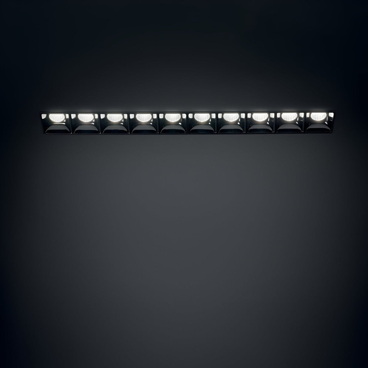 The Phosix Trimless LED Recessed Downlight – 20W, 3000K, Black features a sleek, rectangular design with ten evenly spaced fixtures that offer bright illumination against a dark background. Constructed from durable die-cast aluminum, it ensures a seamless installation that integrates effortlessly into any space.