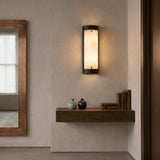 An Elantra LED Small Vertical Alabaster Wall Light in Soft White & Black emits warm light above a floating wooden shelf with two vases and books; a large framed mirror is visible on the left side of this minimalist room.