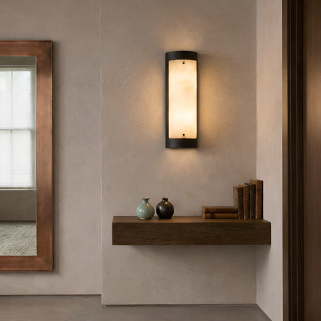 An Elantra LED Small Vertical Alabaster Wall Light in Soft White & Black emits warm light above a floating wooden shelf with two vases and books; a large framed mirror is visible on the left side of this minimalist room.