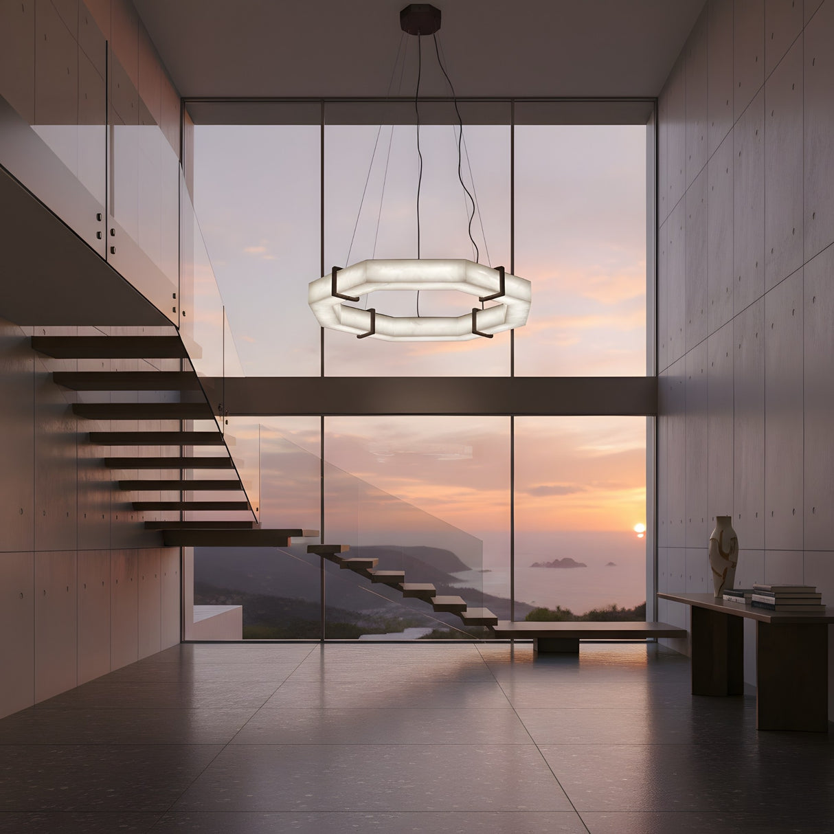A modern, minimalist room with floor-to-ceiling windows overlooks a coastal sunset. Floating stairs and the Vireon LED Small Alabaster Chandelier in Bronze & Soft White enhance the sleek design. A small table with books and a sculpture sits in the corner.