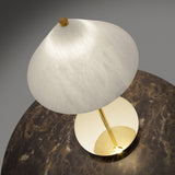 Fuji LED alabaster table lamp with a tilted conical white shade, slim stem, and round polished brass base, shown glowing on a brown marble surface.
