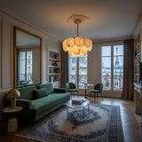 A stylish Parisian living room featuring a green sofa, two green chairs, a rug, large windows with views of the Eiffel Tower and classic architecture, and illuminated by the Solyra 6 Light 2 Tier Petal Alabaster Chandelier in Soft White.