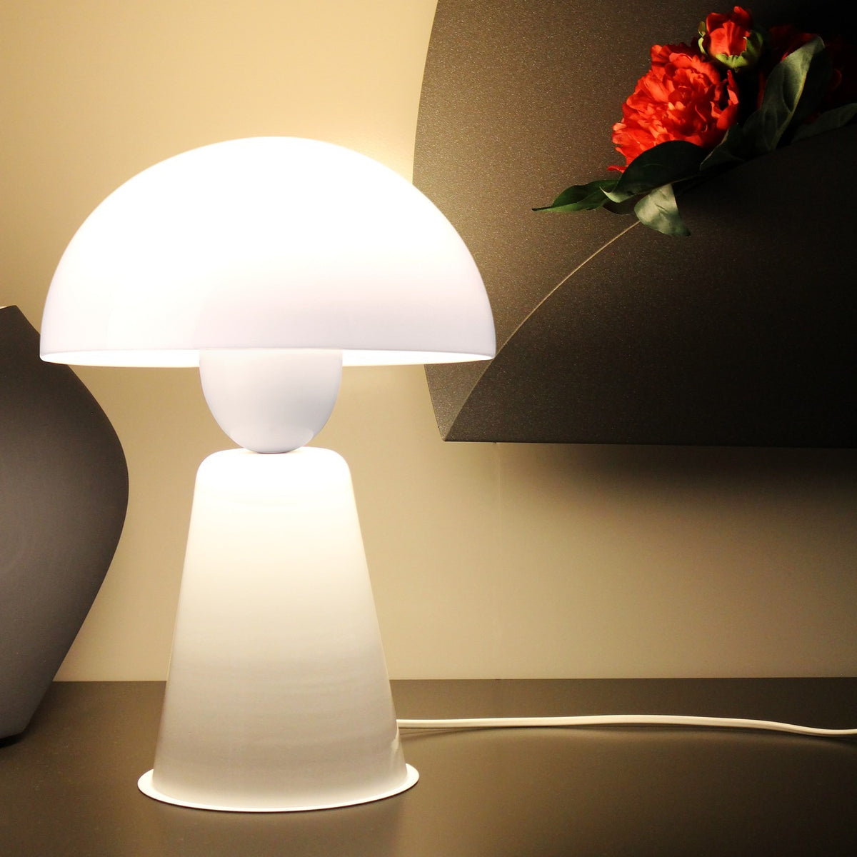 Star Table Lamp in glossy white acrylic and iron, with a dome shade, rounded neck and conical base, lit on a desk beside a dark vase and red flowers.