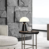Star Table Lamp in glossy black with white acrylic dome shade on a round side table, shown in a living room with sea-view window and stone wall.