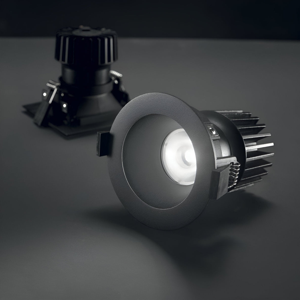 Two Lumos Downlight Round Frame - Black LED ceiling lights are installed on a dark gray surface. One of the downlights is illuminated, casting a bright white glow, while the other remains unlit in the background.