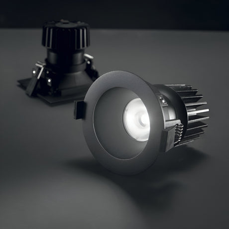 Two Lumos Downlight Round Frame - Black LED ceiling lights are installed on a dark gray surface. One of the downlights is illuminated, casting a bright white glow, while the other remains unlit in the background.