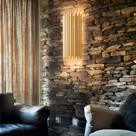 A cozy room with stone walls and two plush armchairs features a modern vertical light fixture that emits a warm glow, highlighting the textured wall. The Fluxor Wall Light in gold enhances the contemporary design, while beige curtains partially cover a window, adding to the inviting atmosphere.