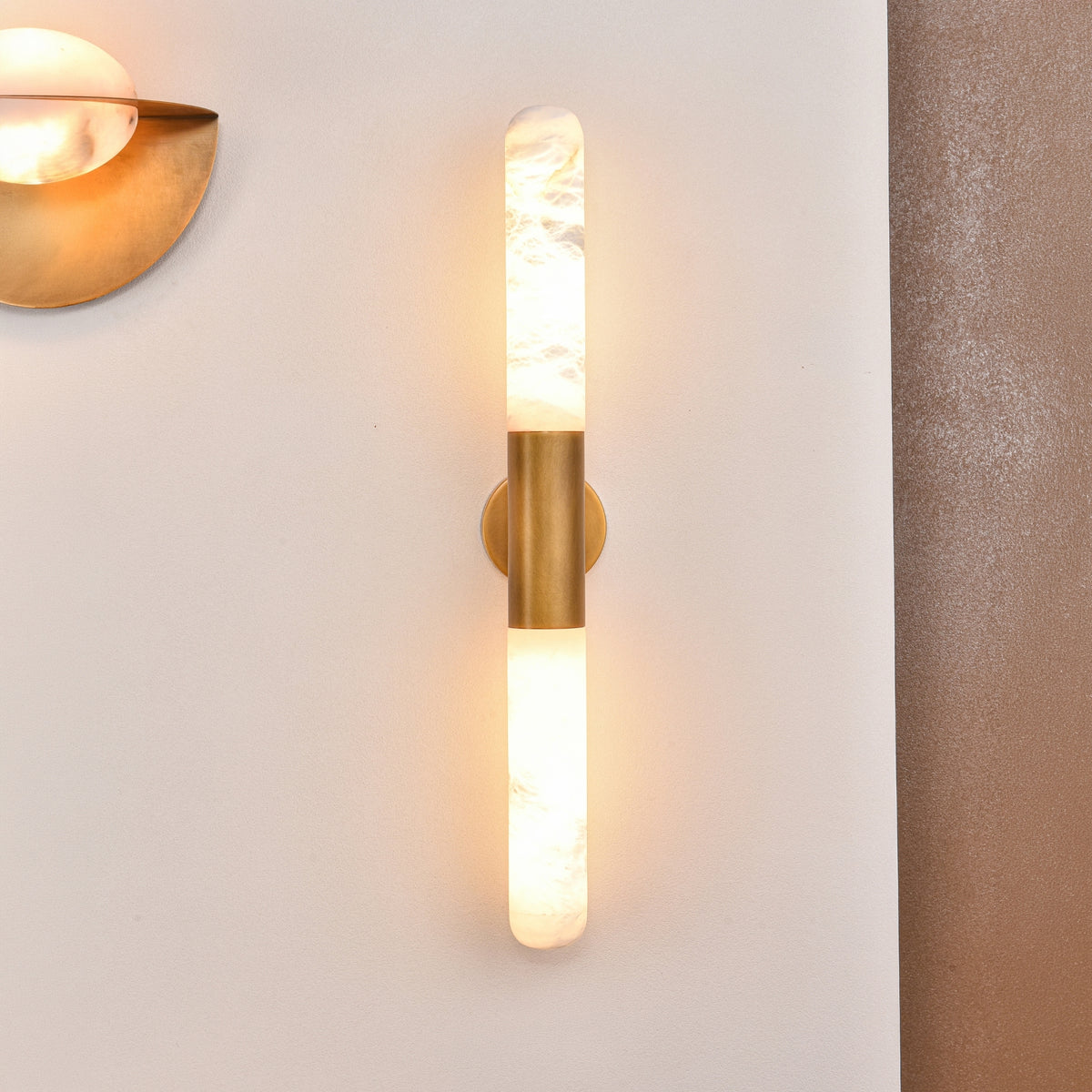 Averin LED Alabaster Wall Light in Brushed Brass, two vertical alabaster columns joined by a brass bracket, glowing warm white on a light wall beside another brass fixture.