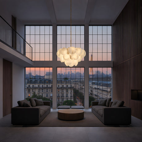 A modern living room with tall windows showing a sunset cityscape, two gray sofas facing a round coffee table, and the Solyra 15 Light 3 Tier Petal Alabaster Chandelier – Soft White with cloud-like lights hanging above.