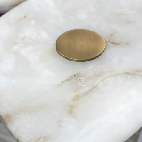 The Calyra LED Alabaster Wall Light in Brass & Soft White, a round flat gold metal fixture, sits atop a smooth white marble surface accented with light beige veining.