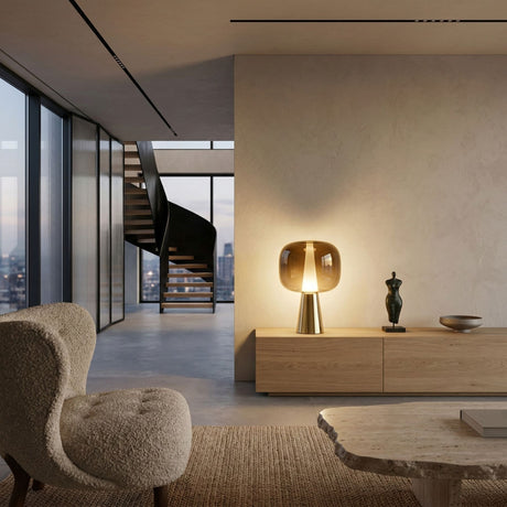 A modern living room with minimalist décor features a cozy textured armchair, a stone coffee table, and a wooden sideboard topped with the Dusk Dawn LED Table Lamp - Gold, plus large city-view windows and a black spiral staircase in the background.