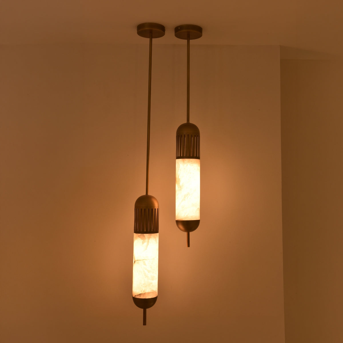 Zerith LED alabaster tube pendant lights in brushed brass, shown as a pair at staggered drop heights, illuminated warm-white in a dimly lit room.