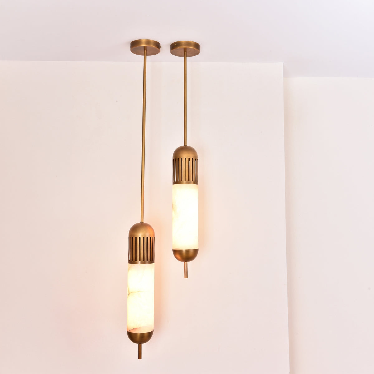 Two Zerith LED alabaster tube pendant lights with brushed brass ceiling mounts and fluted collars, hung at staggered heights against a white wall.