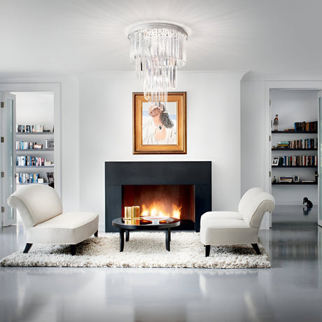 A modern living room featuring a sleek black fireplace is accentuated by two white lounge chairs and a plush white rug, all centered around a round coffee table. The luxury lighting in the room is highlighted by the Emberara 24 Light Crystal Chandelier with a gold finish hanging from the ceiling. A portrait decorates the space above the fireplace, with bookshelves visible in nearby rooms.