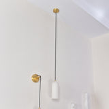 Velmora slim cylindrical alabaster pendant light in soft white, with black flex cable and brass ceiling plate, shown alongside a matching brass wall light in a bright interior.