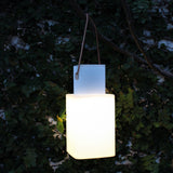 Cuadrat LED Rechargeable Outdoor Table Lamp in white, square glowing shade on a squared base, hung by a leather strap from a tree branch amid dark green foliage.