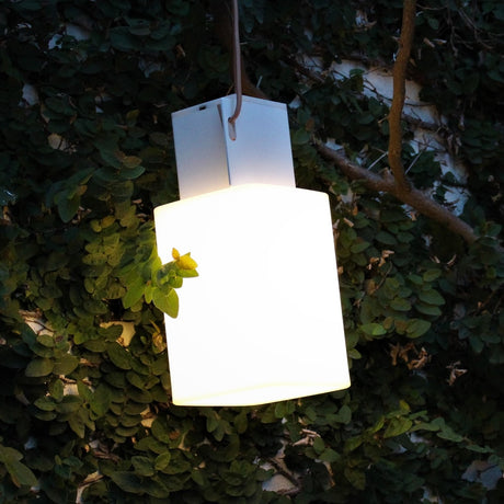 Cuadrat LED rechargeable outdoor table lamp in white, square glowing shade with top handle, shown hanging outdoors amid green foliage at dusk.