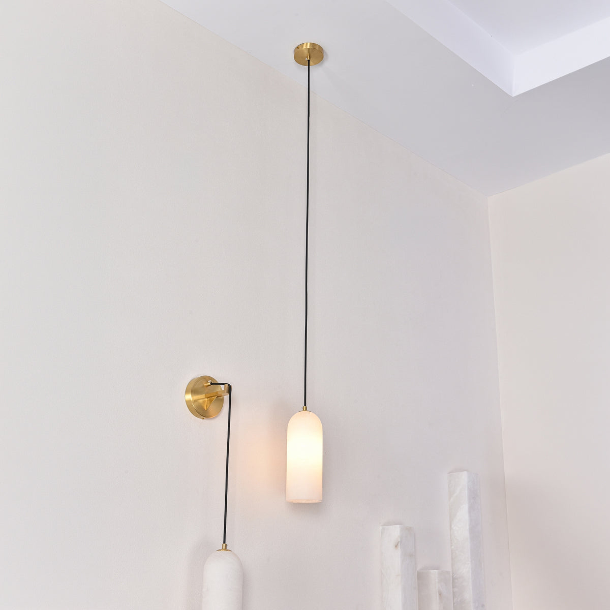 Velmora slim cylindrical alabaster pendant light in soft white, suspended on black flex from a brass ceiling plate, shown illuminated on a wall beside a matching wall light in a neutral room setting.