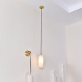 Velmora slim cylindrical alabaster pendant light in soft white, suspended on black flex from a brass ceiling plate, shown illuminated on a wall beside a matching wall light in a neutral room setting.