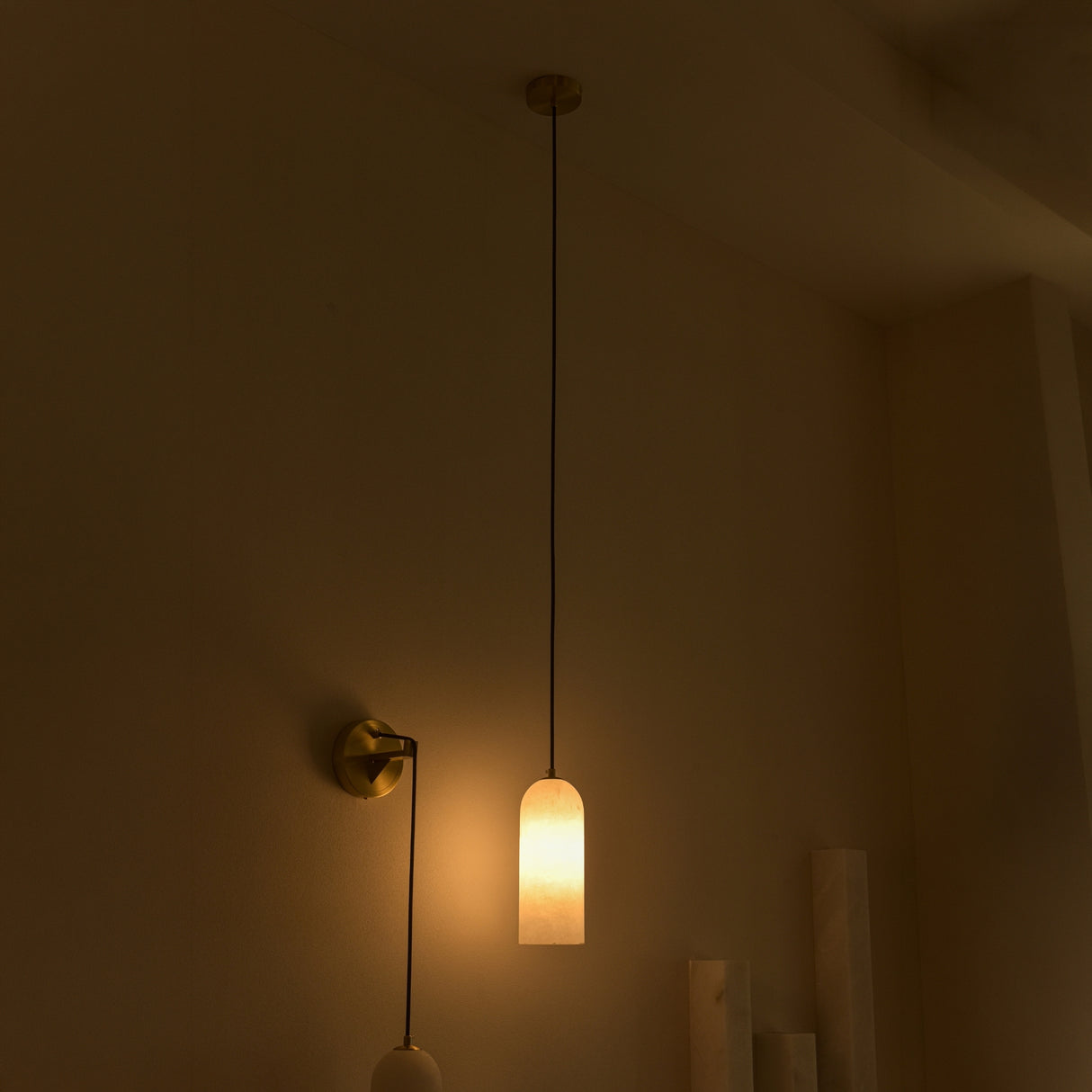 Velmora slim cylindrical alabaster pendant light with brass ceiling plate and black flex, glowing warmly in a dimly lit room beside a wall-mounted brass sconce.