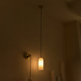 Velmora slim cylindrical alabaster pendant light with brass ceiling plate and black flex, glowing warmly in a dimly lit room beside a wall-mounted brass sconce.