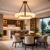 A modern dining room features a round table, six upholstered chairs, and an open kitchen with dark cabinets and marble surfaces. The Cintura LED Medium Alabaster Suspension Light in soft white & brown hangs under floor-to-ceiling windows overlooking greenery.
