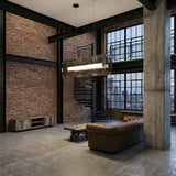 Niori Qbic LED Large Linear Suspension Light in clear glass with brass corners hangs in an industrial loft above a leather sofa near a spiral staircase.