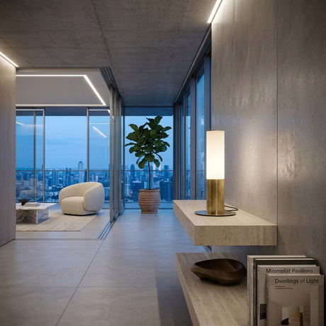 Modern apartment interior at dusk with minimalist decor, potted plant, white armchair, and large windows showing the city skyline. In the foreground, shelves display design books and the Magari Table Lamp - Gold.