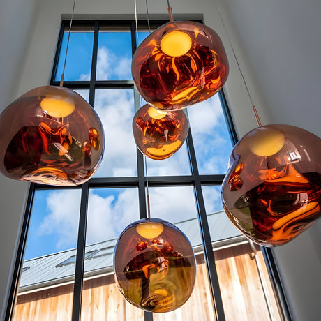 Several Lava Medium LED Pendant Lights in copper hang from the ceiling before tall windows, revealing blue skies and clouds outside with a wooden building visible below.