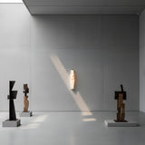Minimalist gallery with tall gray walls, three abstract geometric sculptures on pedestals, and the Aurelia LED Alabaster Wall Light – Soft White illuminated by natural sunlight from a skylight.