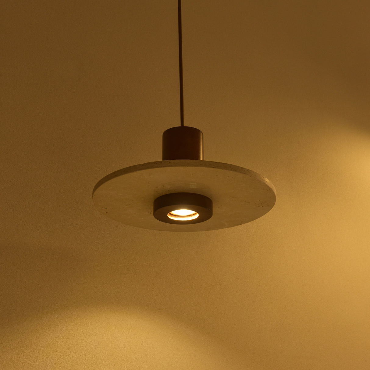 Torvane 1 Light Travertine Disc Pendant Light with light beige stone disc, dark walnut brown wooden column, and recessed downward-facing light, shown illuminated against a warm wall.