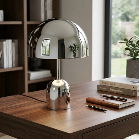 The Rosa Table Lamp - Chrome sits on a wooden desk with notebooks and pens. Bookshelves and a large window with greenery are in the background, while the lamp’s chrome finish reflects the room and window.