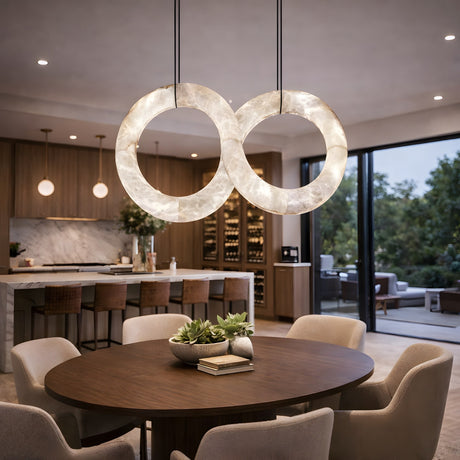 A modern dining area with a round wooden table, beige chairs, and a centerpiece bowl is illuminated by the Serava LED Alabaster Loop Suspension Light – Soft White. In the background, the kitchen features large windows framing lush greenery.