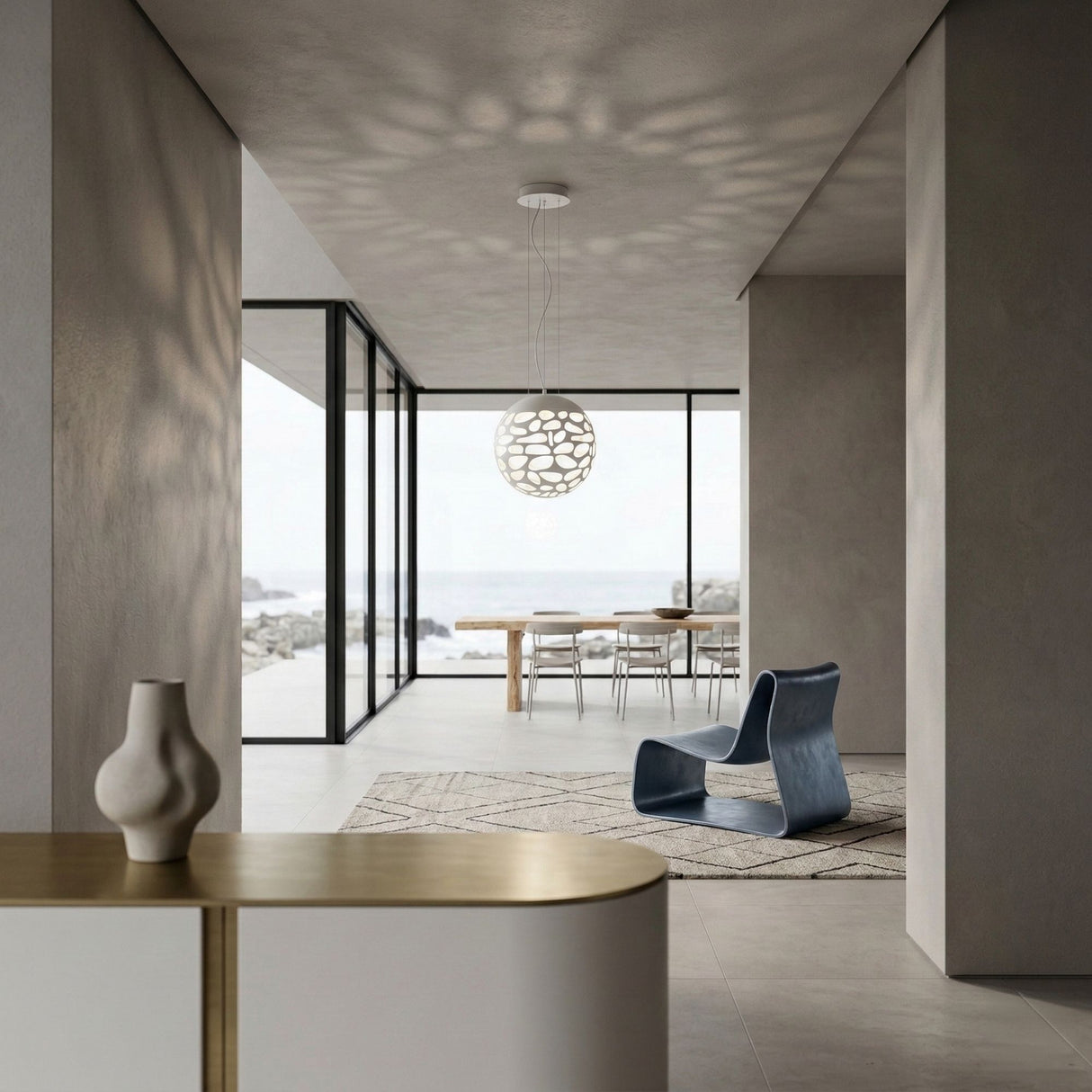 Minimalist living space with large glass windows overlooking a rocky shoreline, modern dining table and chairs, the Elon LED Light Pendant in Matte White overhead, blue sculptural chair, neutral walls, and a decorative vase on the sideboard.