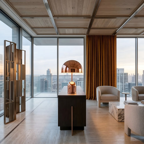 Modern living room with floor-to-ceiling windows overlooking a cityscape, light wood floors and ceiling, beige chairs, a marble coffee table, the Rosa Table Lamp - Copper on a dark console, and orange curtains.