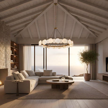 A modern living room with a vaulted white wood ceiling features the Solaryn LED Linear Disc Alabaster Chandelier in Soft White & Brass, a stone fireplace, light sectional sofa, round coffee table, and floor-to-ceiling windows with sunset ocean views.
