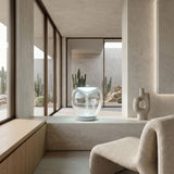 A modern, minimalist room in neutral tones features the Empatia LED Table Lamp - Clear, a sculptural, energy-efficient hand-blown glass lamp on a ledge near large windows overlooking cacti and rocks, with a textured chair and decorative vase nearby.