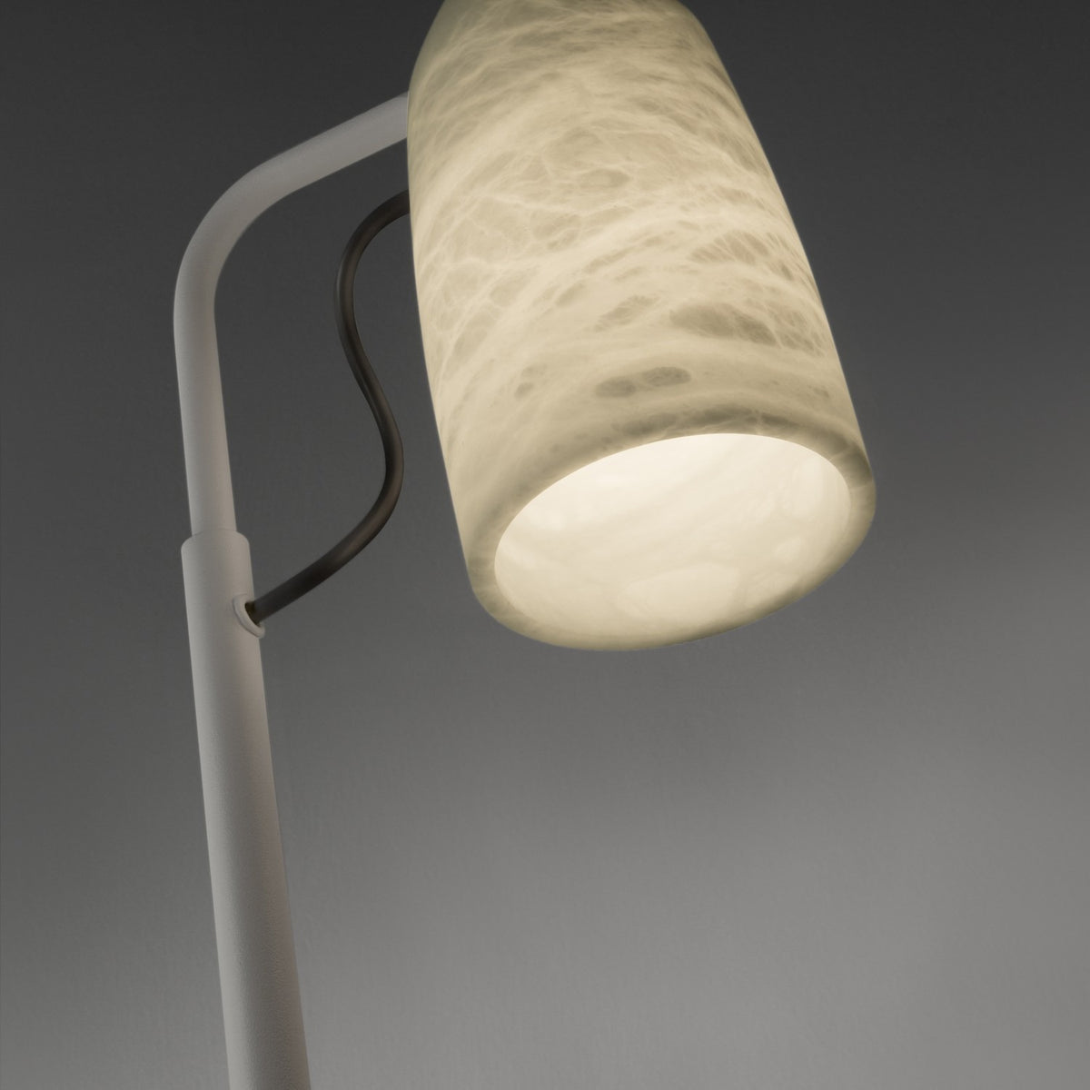 Close-up of the Paros Alabaster Table Lamp's cylindrical alabaster shade with soft grey veining, glowing downward from a white iron arm and stem against a grey background.