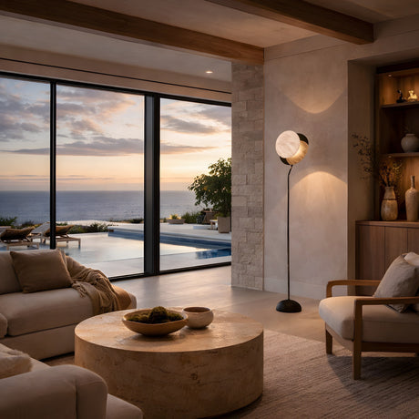 A cozy living room with neutral decor overlooks a pool and ocean at sunset through large glass doors, featuring soft seating, a round stone coffee table, and the Selvano 1 Light Globe Alabaster Floor Lamp in Soft White & Matte Black.