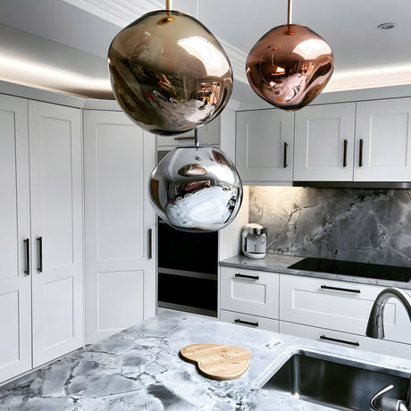 Modern kitchen with white cabinets, marble-patterned countertops and backsplash, ambient lighting from a Lava Medium LED Pendant Light - Gold, a sleek sink, and a heart-shaped wooden cutting board near the sink.