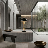 A modern minimalist interior with concrete walls and floors features the Ascot 4 Light Large Table Lamp in black & gold. Glass doors open to a courtyard tree, while bronze bowls and contemporary furniture complete the scene.
